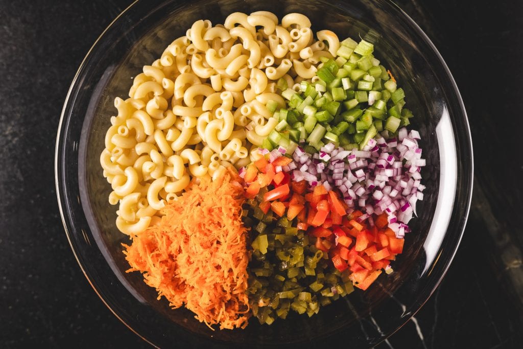 Cooked macaroni noodles in a bowl, topped with shedded carrots, diced pickles, diced red onion, diced bell pepper, and diced celery.