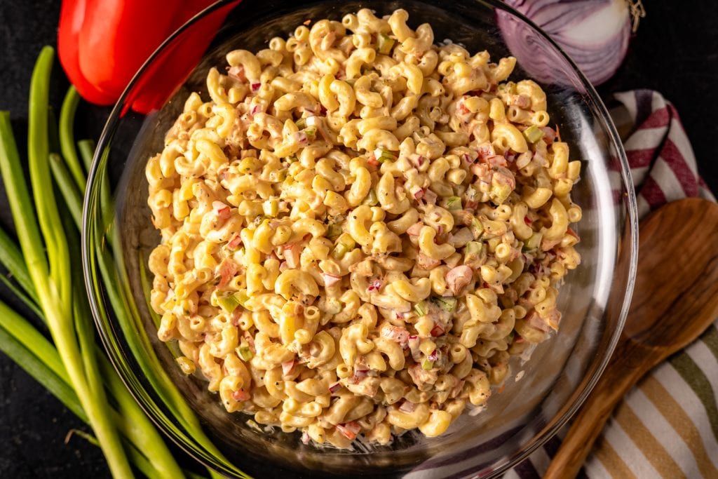 Macaroni salad in a bowl, surrounded by a striped dish towel and wooden spoon.
