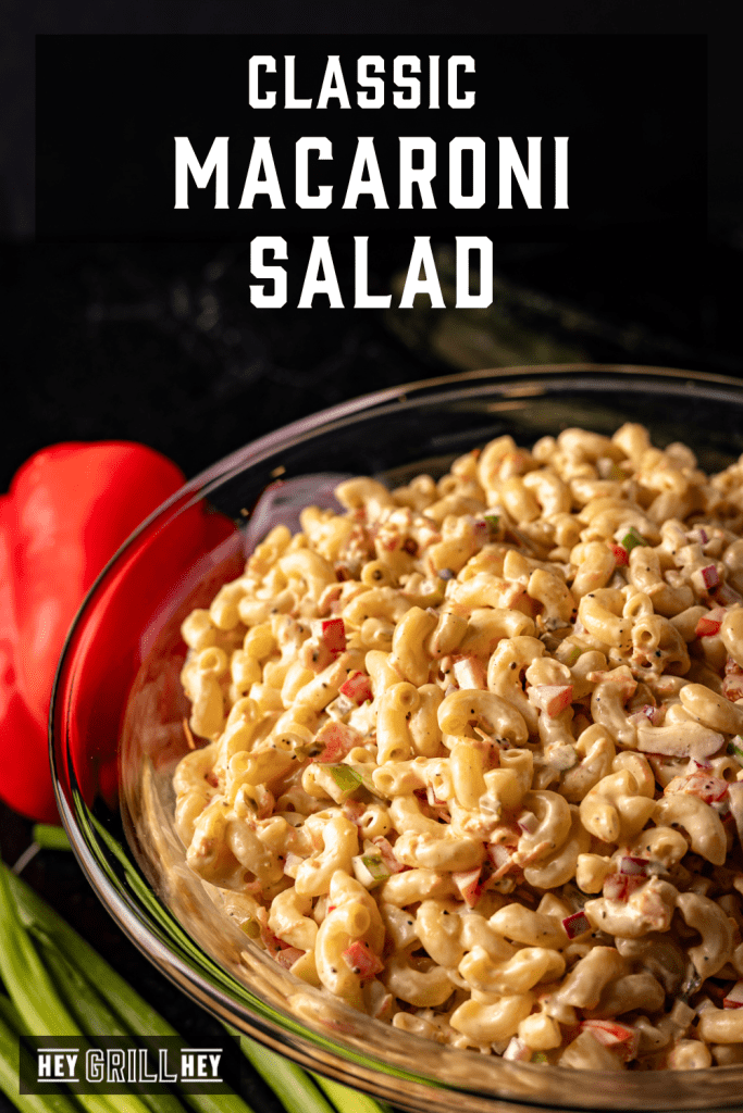 A glass bowl filled with macaroni salad. A red bell pepper and green onions are near the bowl.