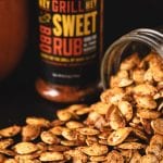 Smoked pumpkin seeds spilling out of mason jar in front of container of Hey Grill Hey Sweet Rub.