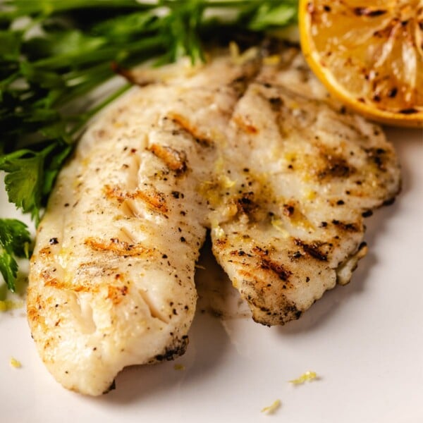 Grilled tilapia with lemon wedge and parsley leaves on white plate.