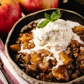 Apple crisp in white bowl topped with cream, in front of whole apples.