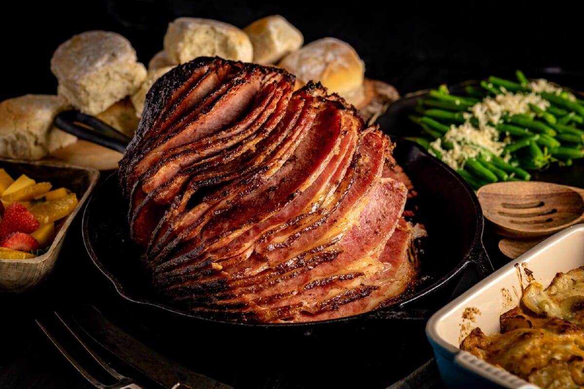 Sliced ham in skillet surrounded by biscuit, asparagus, and side dishes.