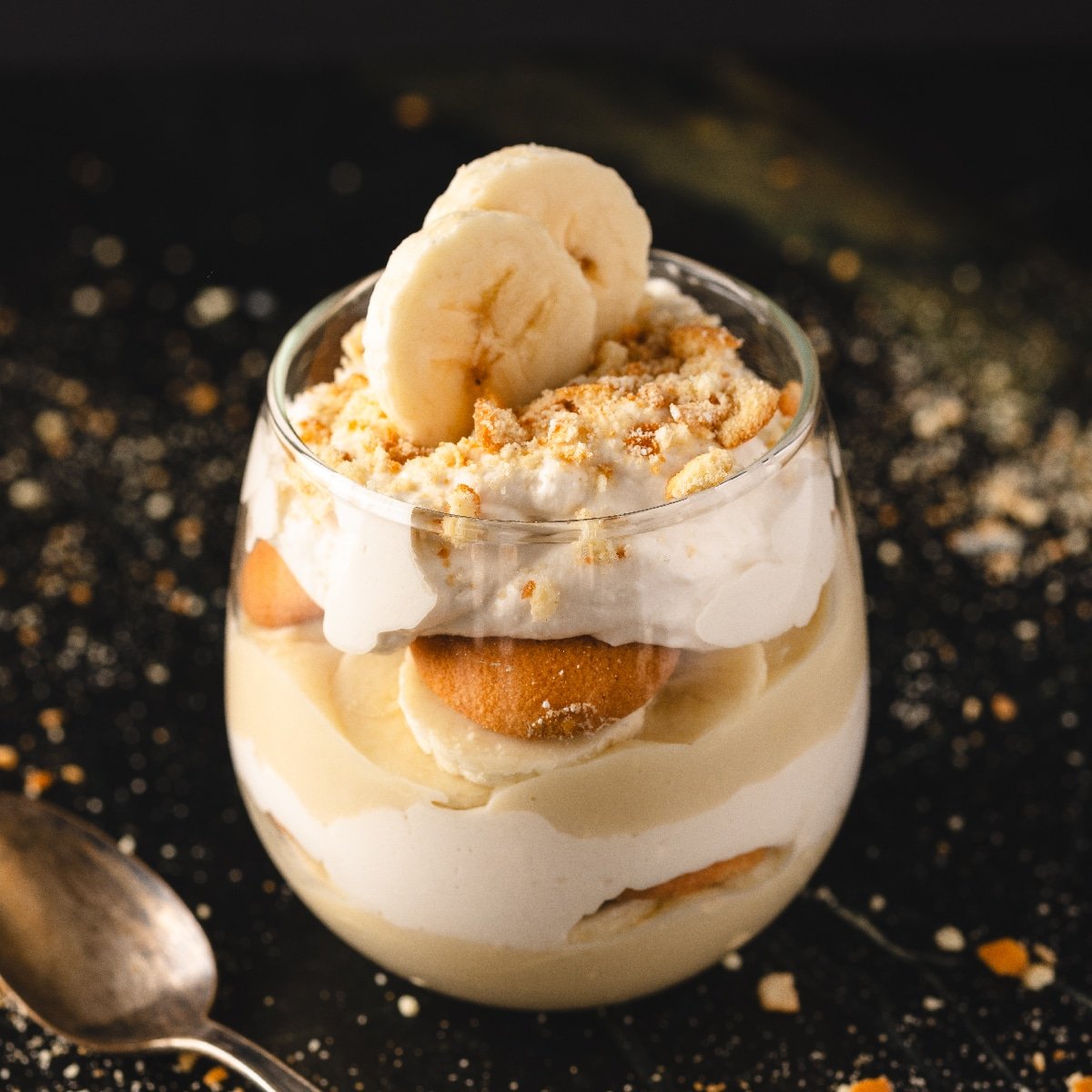 Banana pudding in stemless wineglass next to spoon and wafer crumbles.