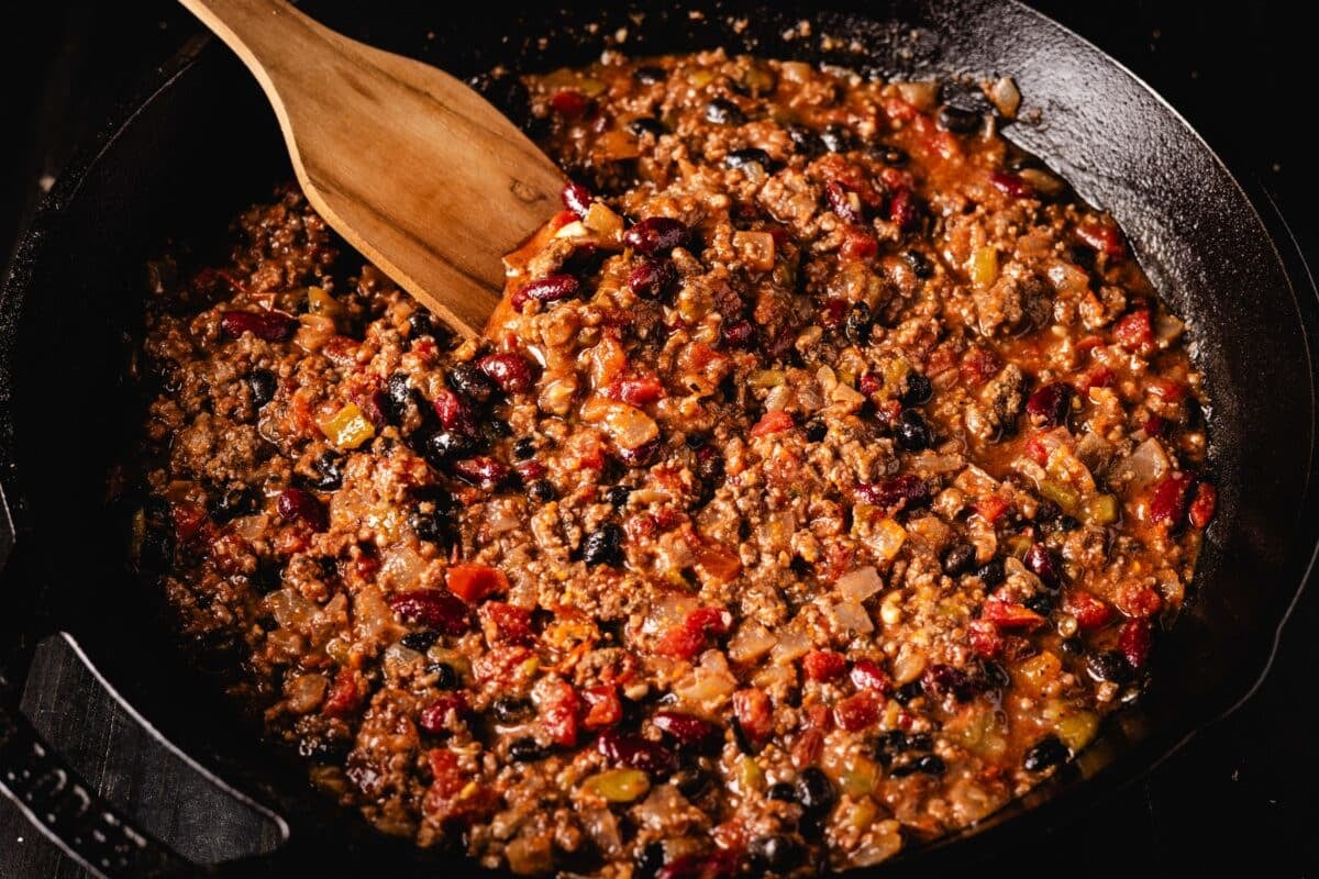 A wooden spatula stirring ground beef chili in a skillet.