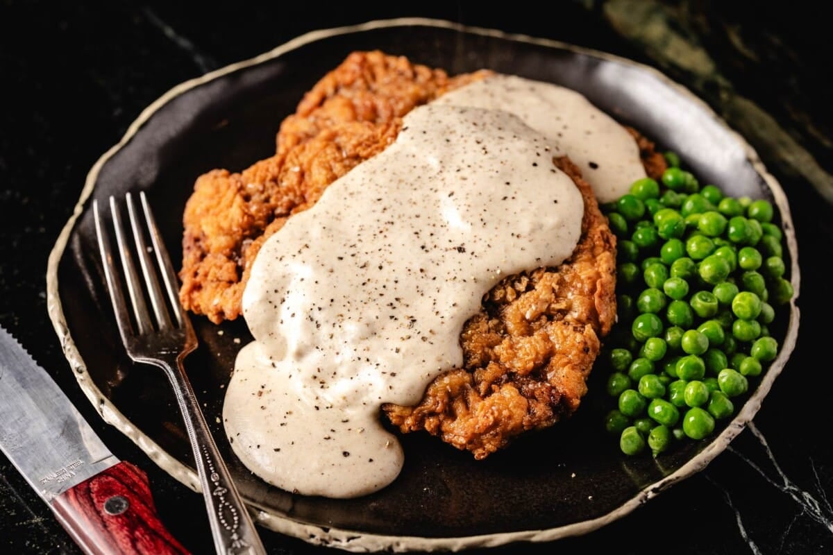 Fried steak with gravy on plate with peas, and fork.