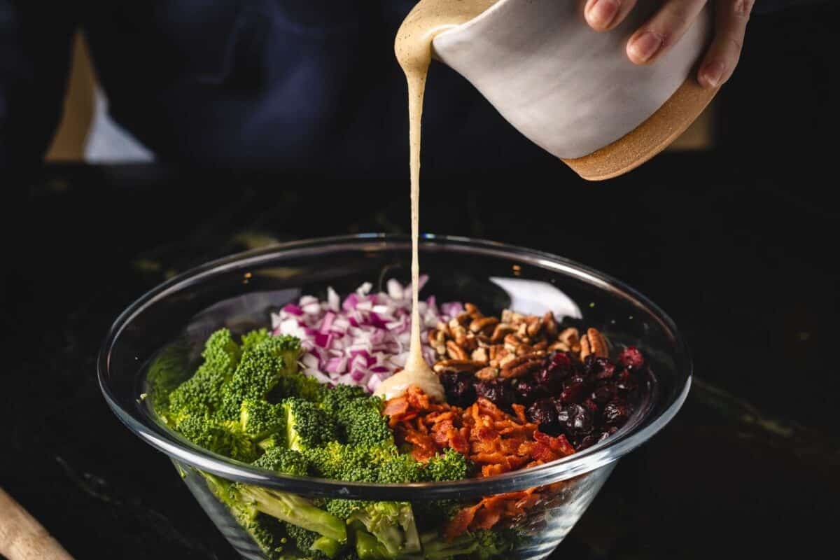 Dressing in cup pouring into veggie mix in glass bowl.