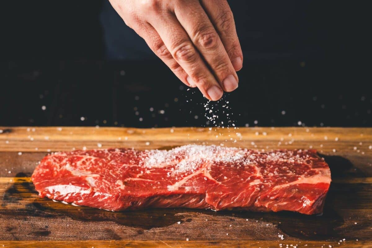 Flat iron steak on cutting board being seasoned with Kosher salt.