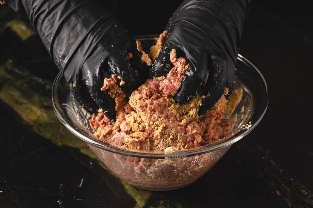 Gloved hands mixing beef mixture in glass bowl.