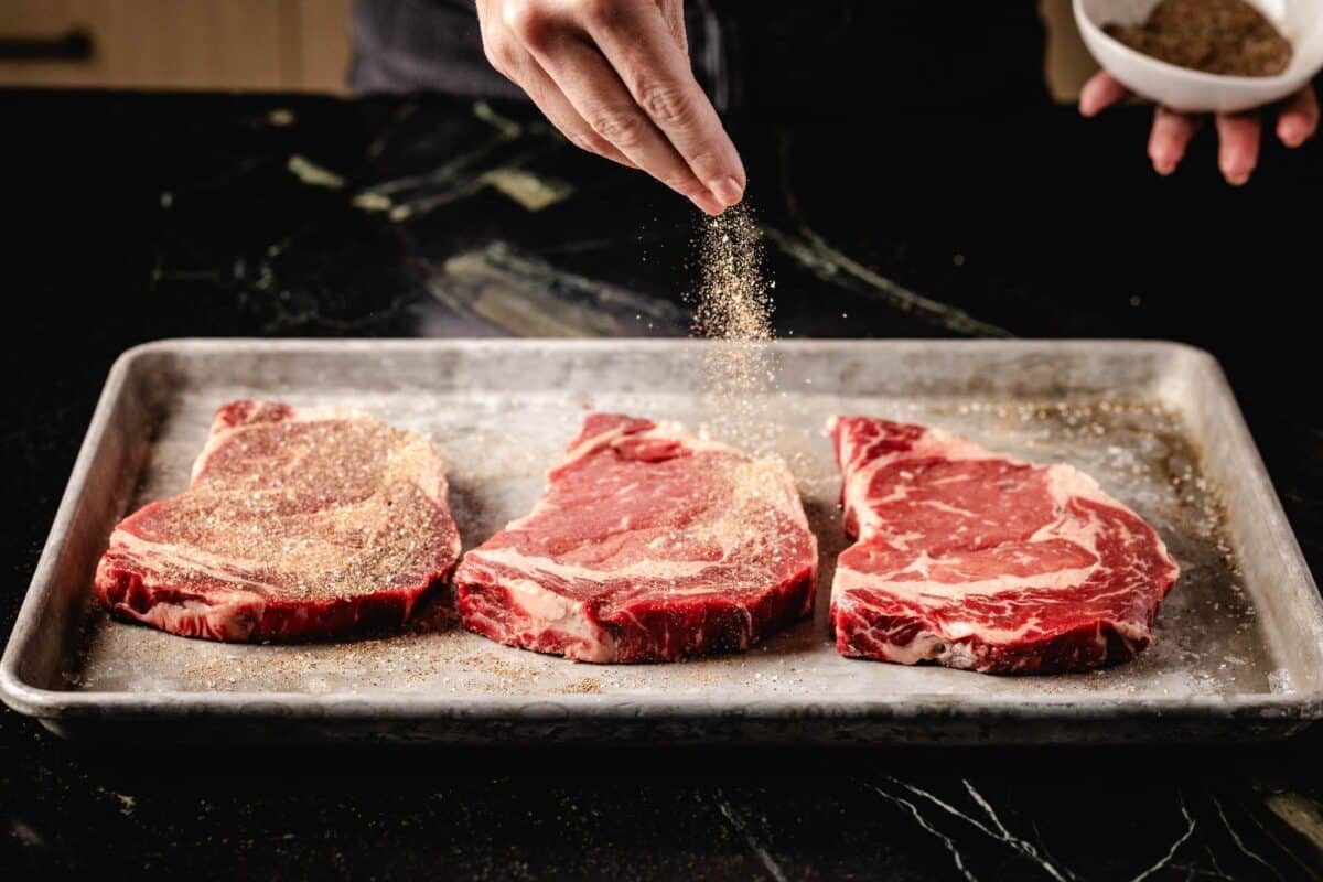 Raw steaks on metal pan sprinkled with seasoning.