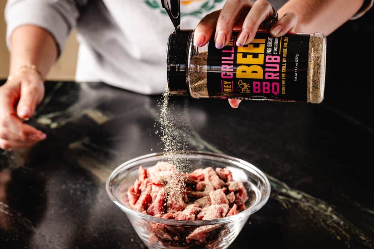 Raw steak in glass bowl being seasoned with Beef Rub.