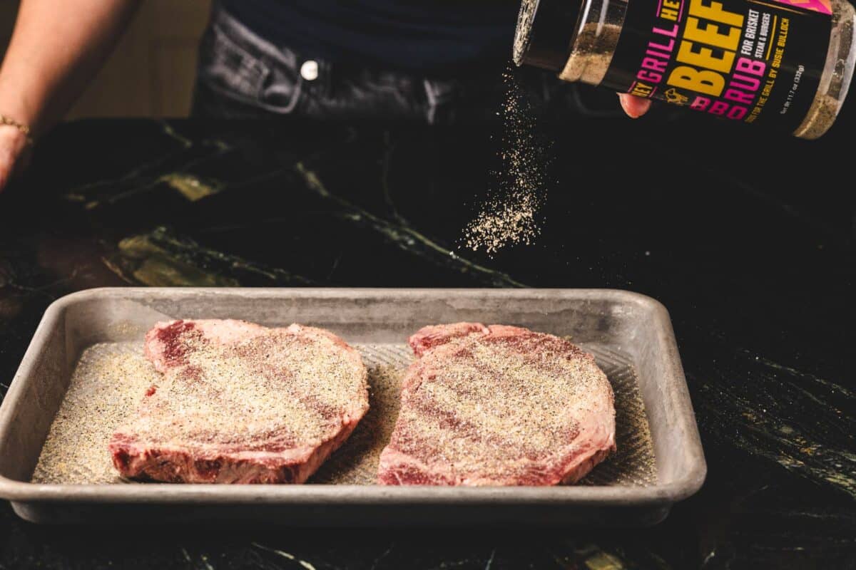 Ribeye steaks on baking sheet being seasoned with Beef Rub.