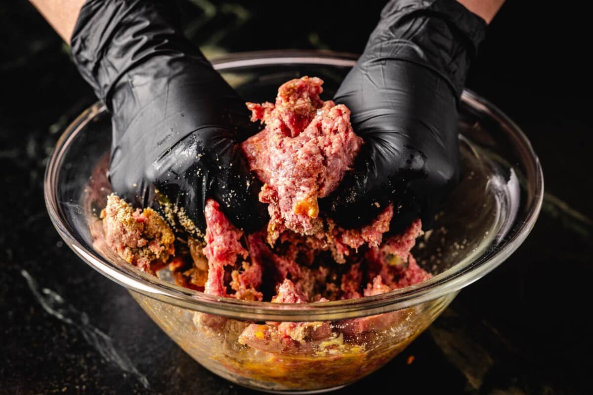 Ground beef in glass bowl being mixed by hands wearing black gloves.