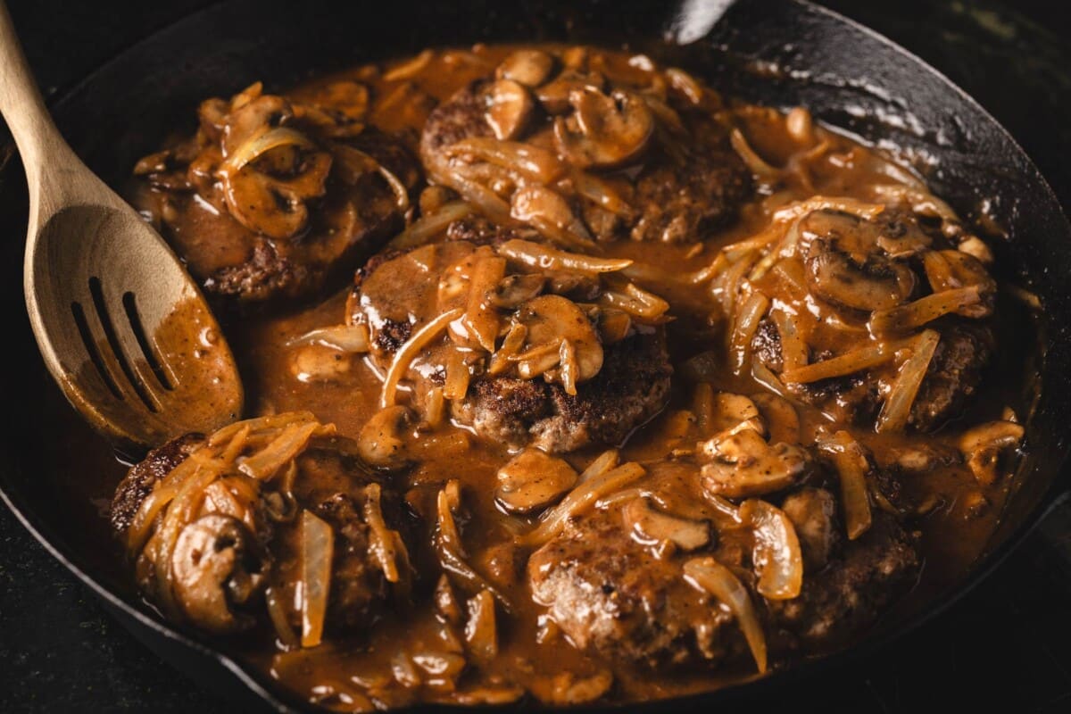Salisbury steaks in mushroom gravy in skillet with wooden spoon.