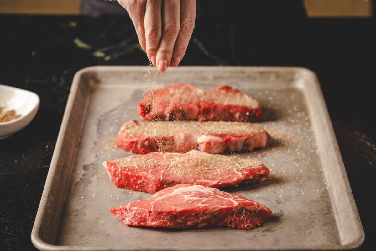 Raw beef steaks on baking sheet sprinkled with seasoning.