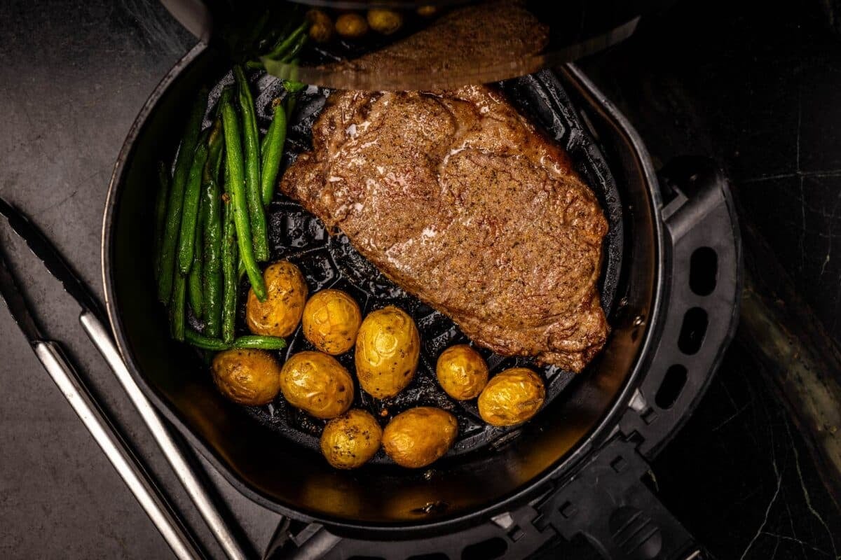 Steak, potatoes, and green beans in air fryer tray.