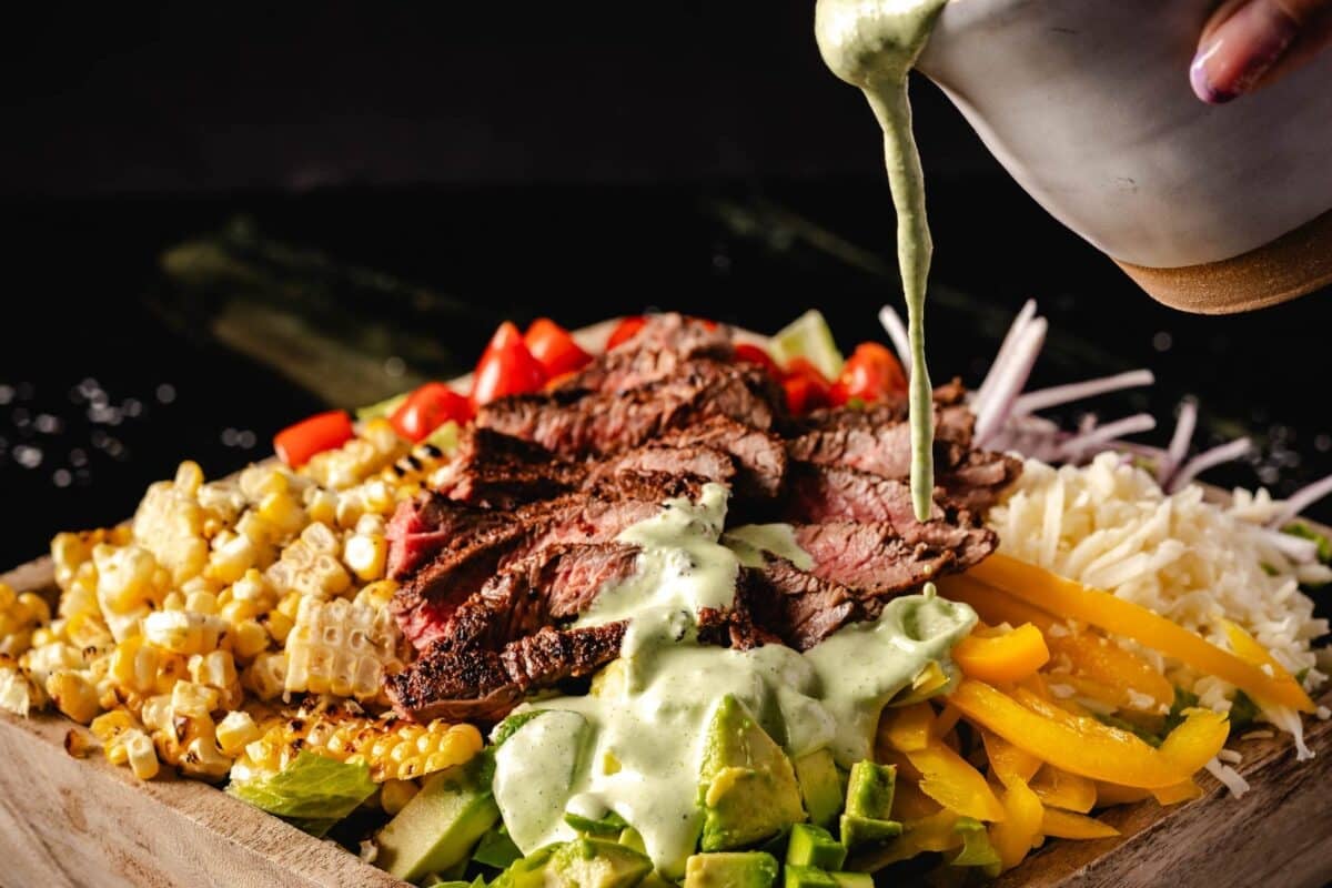 Steak salad ingredients in bowl being drizzled with jalapeño lime dressing.