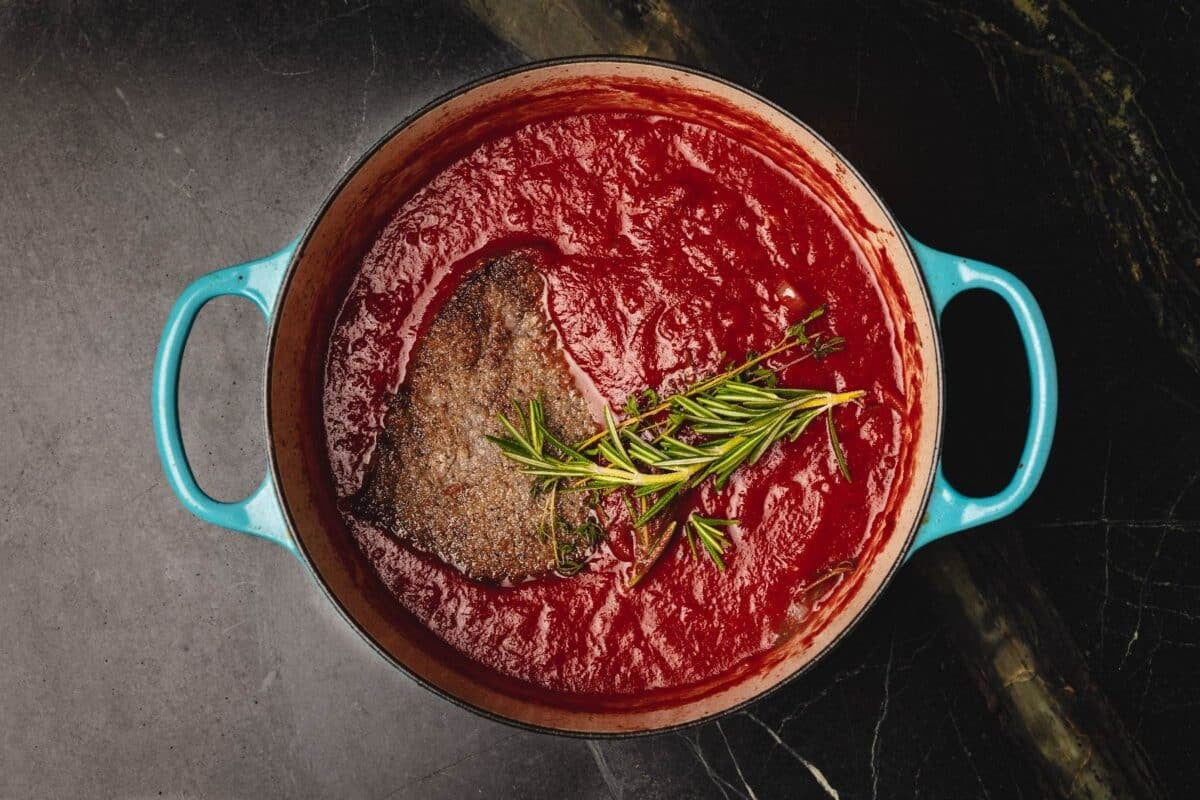 Steak in tomato braise with thyme sprig, in large pot.