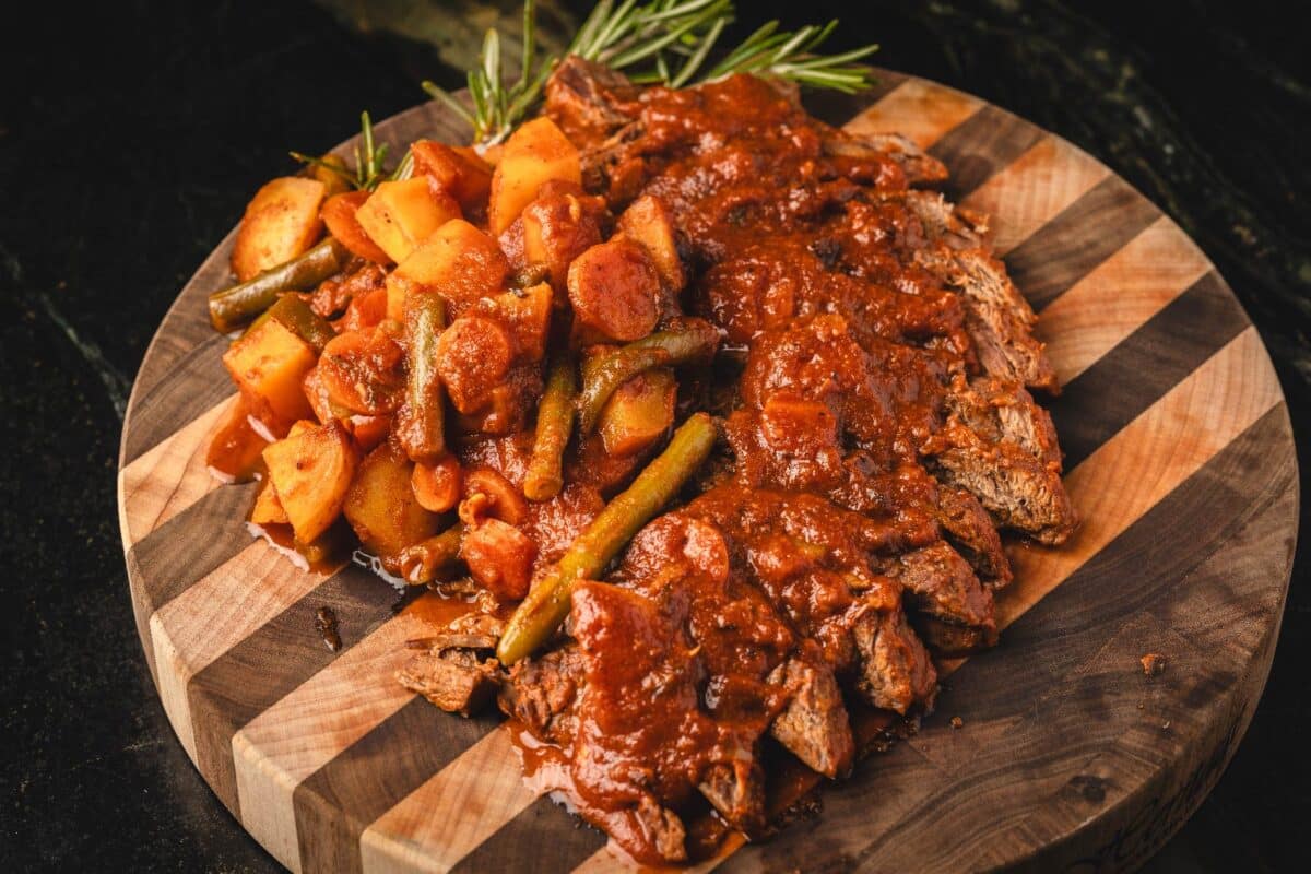 Sliced Swiss steak topped with tomato sauce and carrots, on wooden platter.