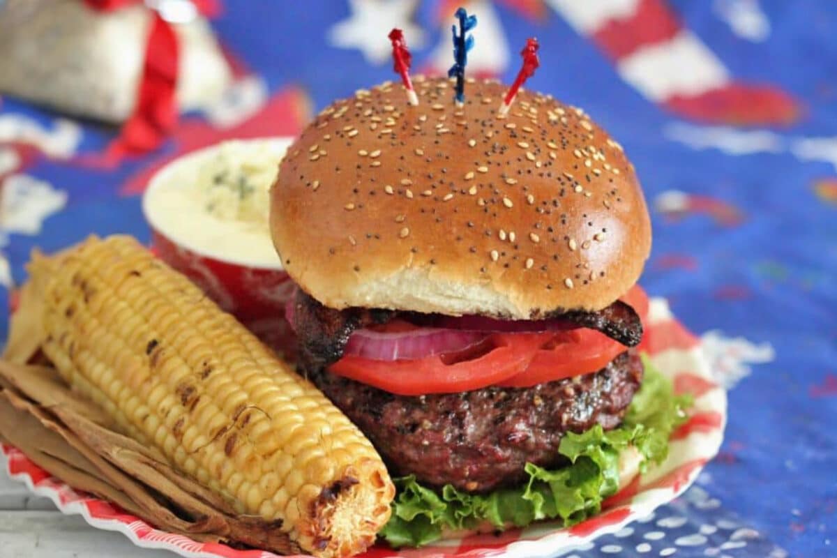 Burger and grilled corn on plate on patriotic tablecloth.