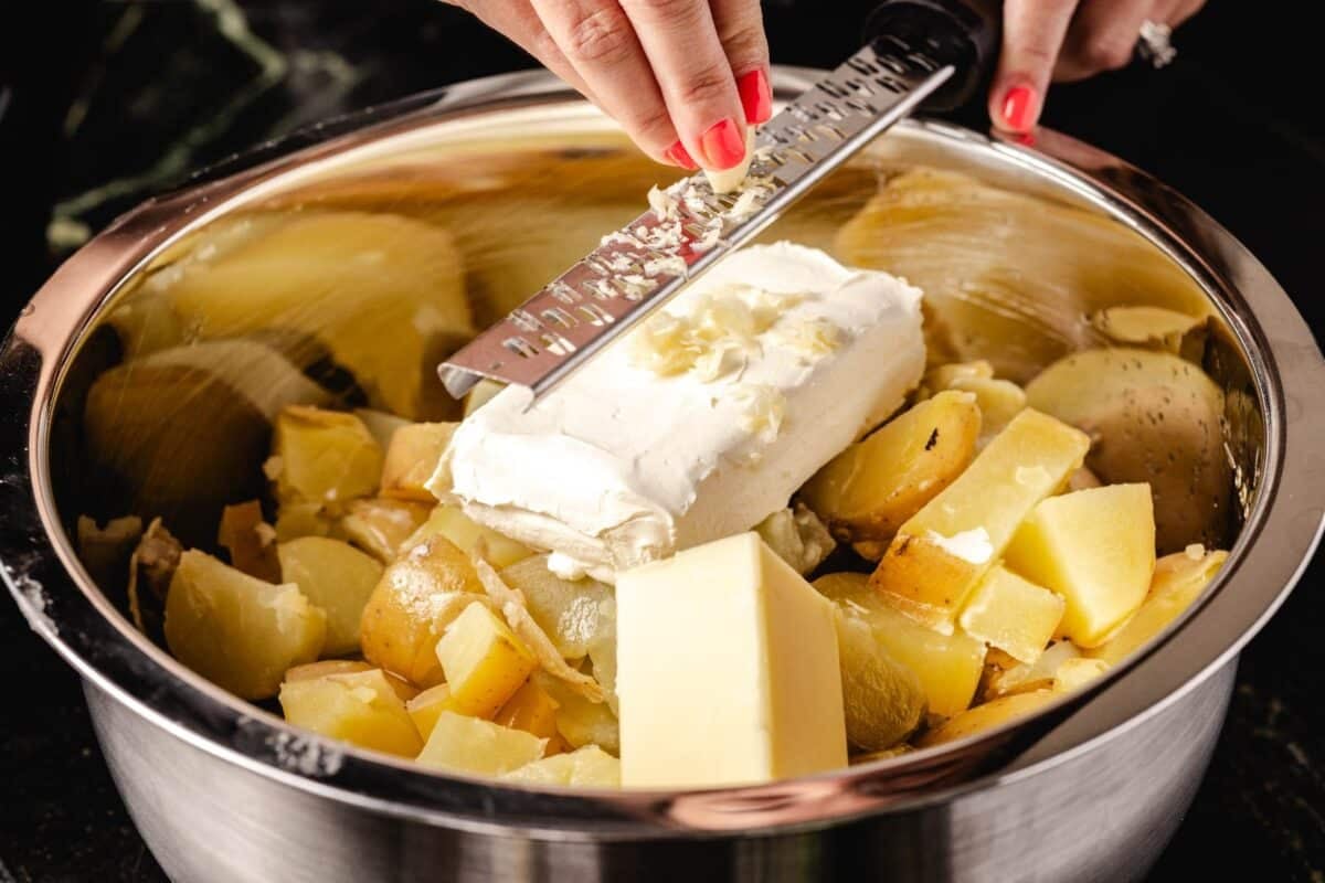 Butter, cream cheese, and potatoes in pot with garlic being shredded onto it.