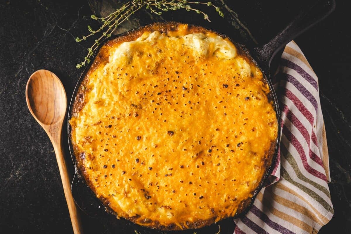 Baked cottage pie in skillet next to wooden spoon.