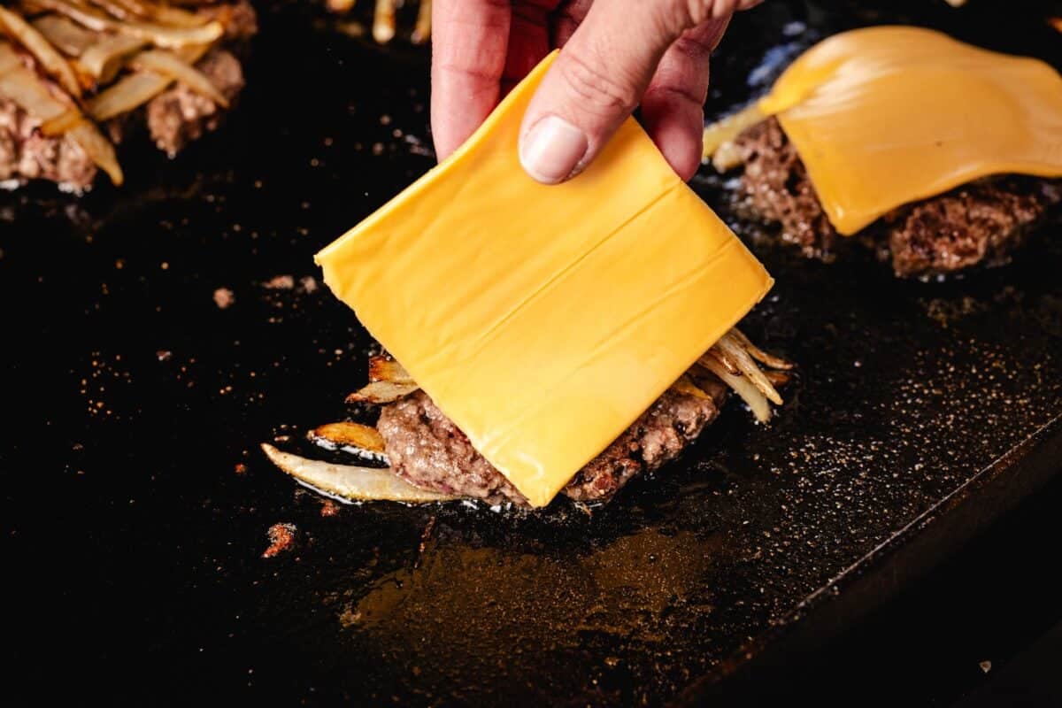 A hand placing cheese slice on smash burger patties.
