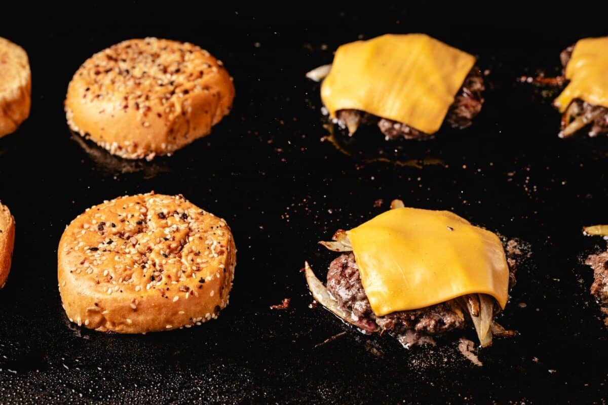 Burger buns and patties with cheese on flat top grill.