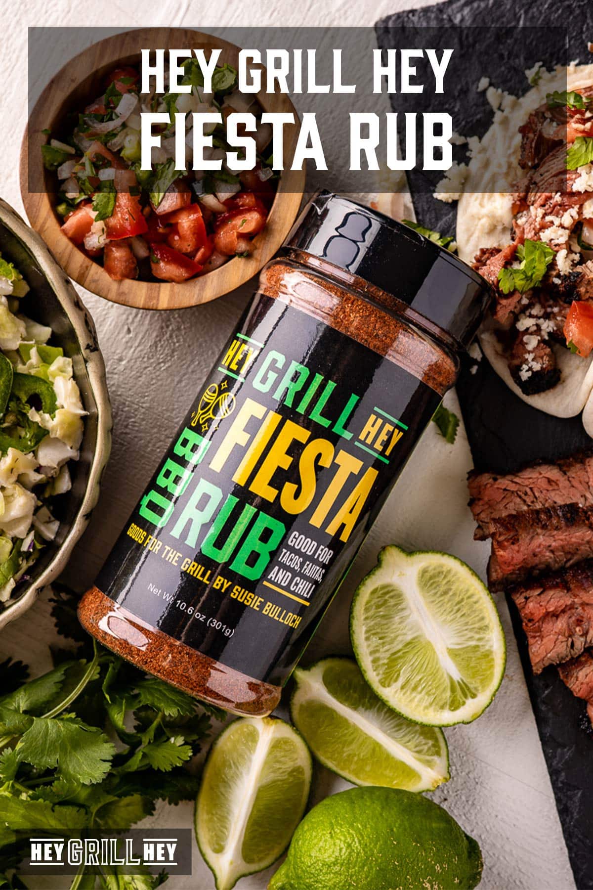 A bottle of rub next to lime wedges and bowls of condiments. Text reads "Hey Grill Hey Fiesta Rub".