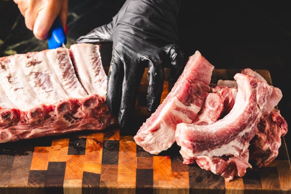 Rack of baby back ribs on cutting board being sliced with knife, and held with gloved hand.