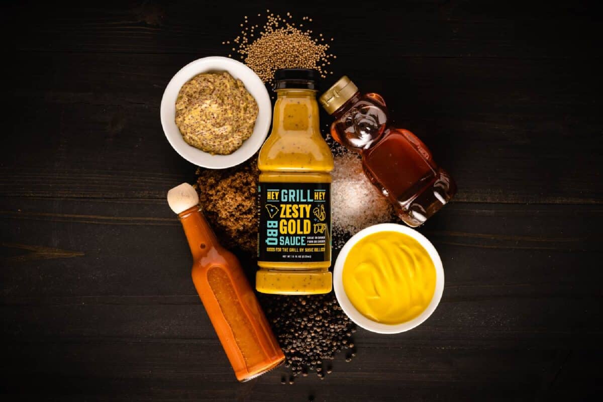 Bottle of Zesty Gold BBQ Sauce surrounded by raw ingredients on black tabletop.