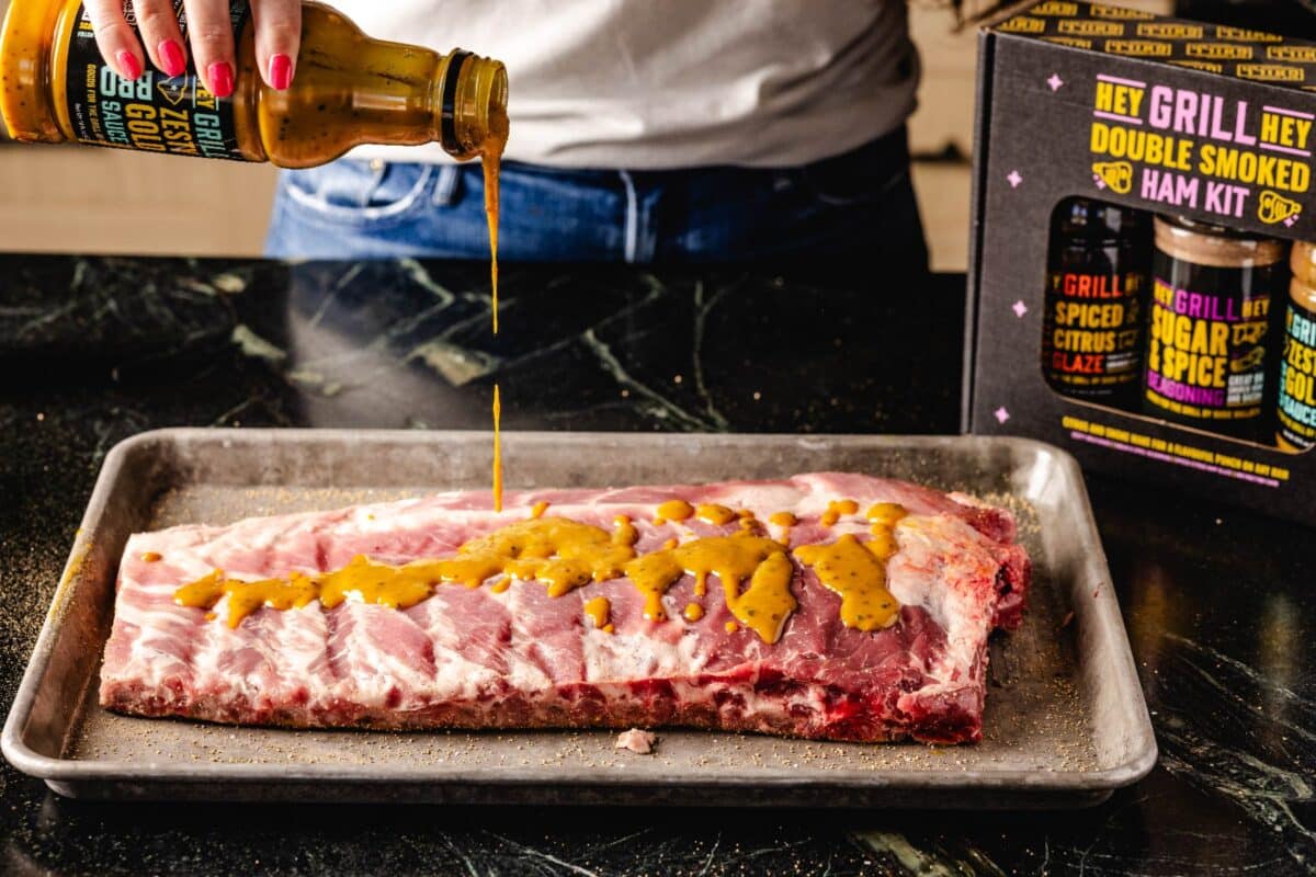 Zesty Gold sauce being drizzled on a rack of ribs, in front of the Double Smoked Ham Kit.