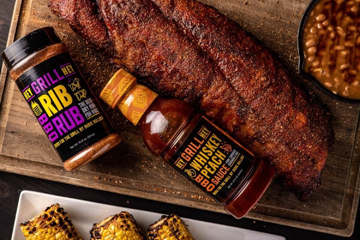 Bottle of sauce and bottle of rub with rack of ribs and sides on serving platter.