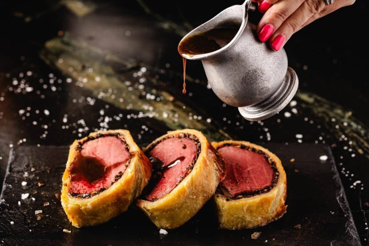 Sliced Beef Wellington on cutting board being drizzled with red win sauce in metal cup.