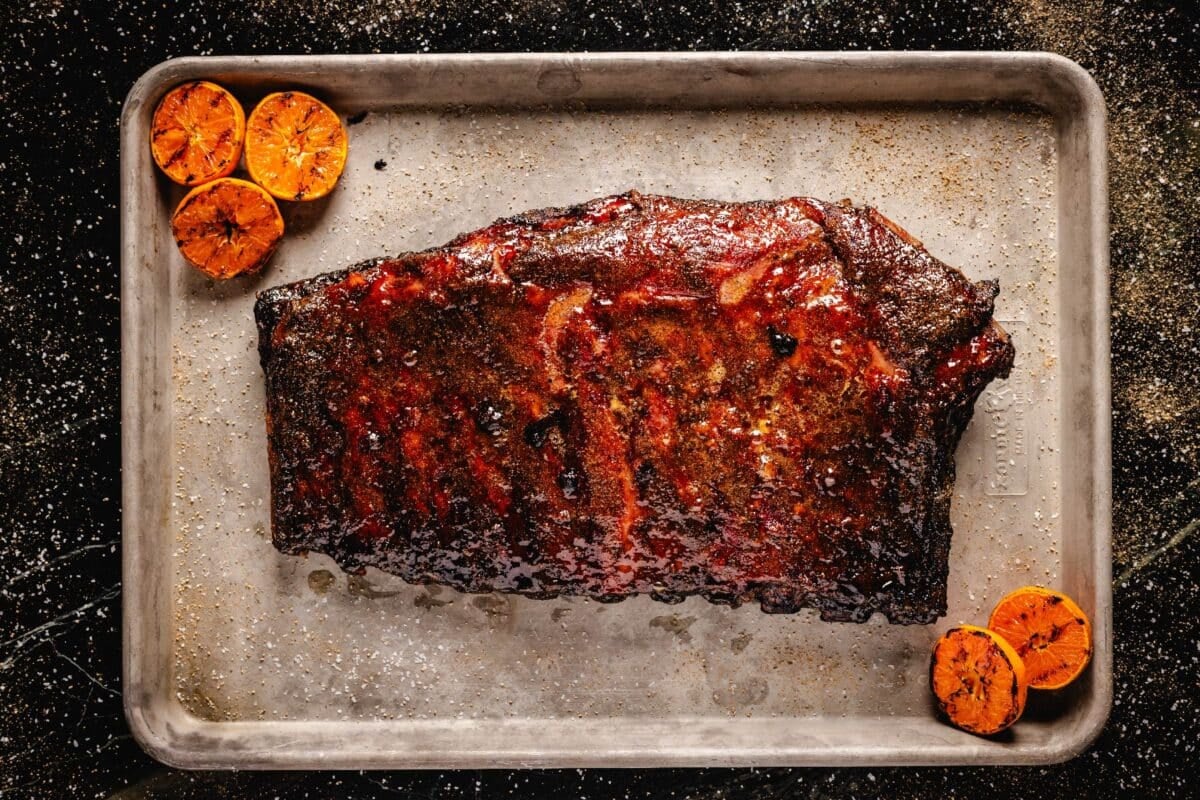 Smoked, unsliced rib rack on a baking sheet with halved oranges.