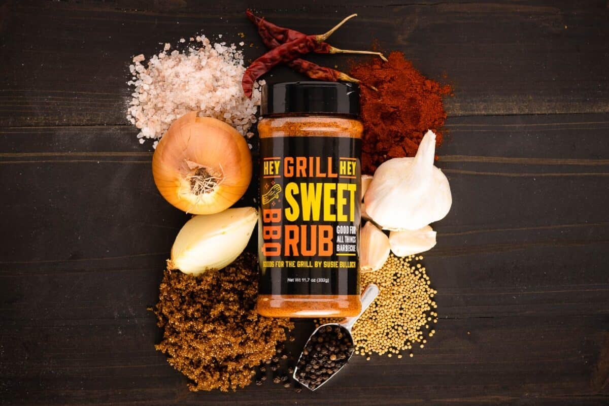 Bottle of Sweet rub on black surface surrounded by raw seasoning ingredients.