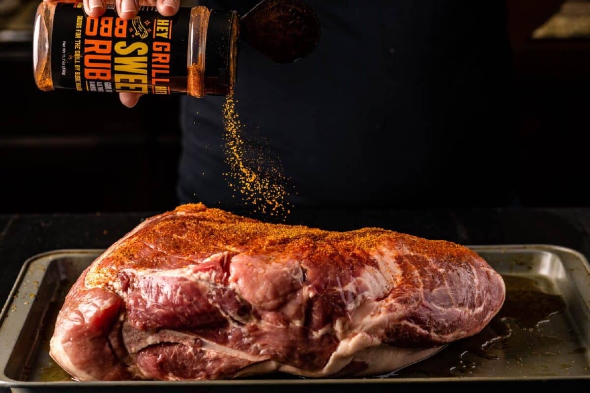 Pork shoulder on baking sheet being sprinkled with seasoning.