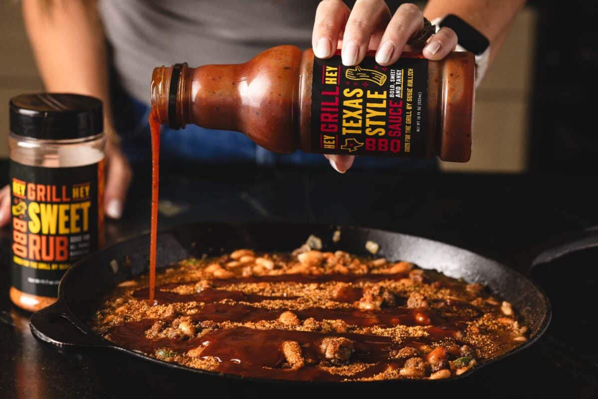 Beans in skillet with Texas Style BBQ Sauce being poured in.