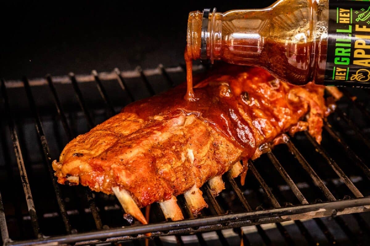 A rack of ribs on the smoker being drizzled with sauce.