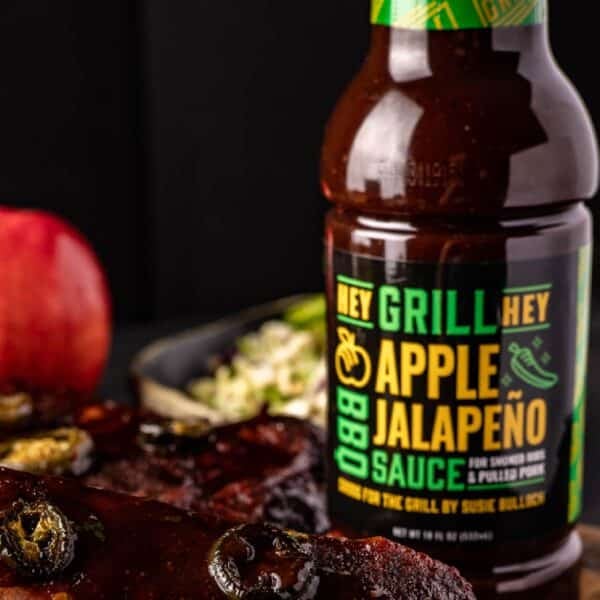 A bottle of Apple Jalapeño BBQ Sauce on countertop next to apples and other raw ingredients.