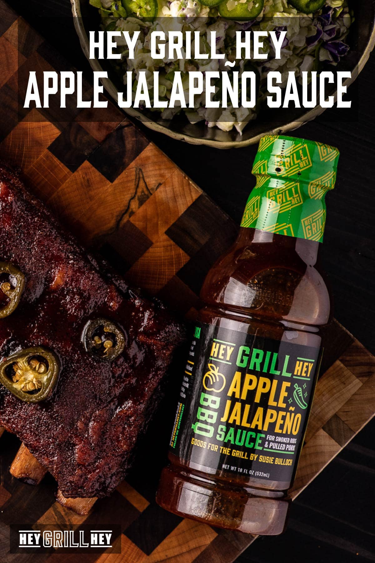 A bottle of BBQ sauce on a cutting board next to a rack of ribs and a bowl of salad. Text reads "Hey Grill Hey Apple Jalapeño Sauce."