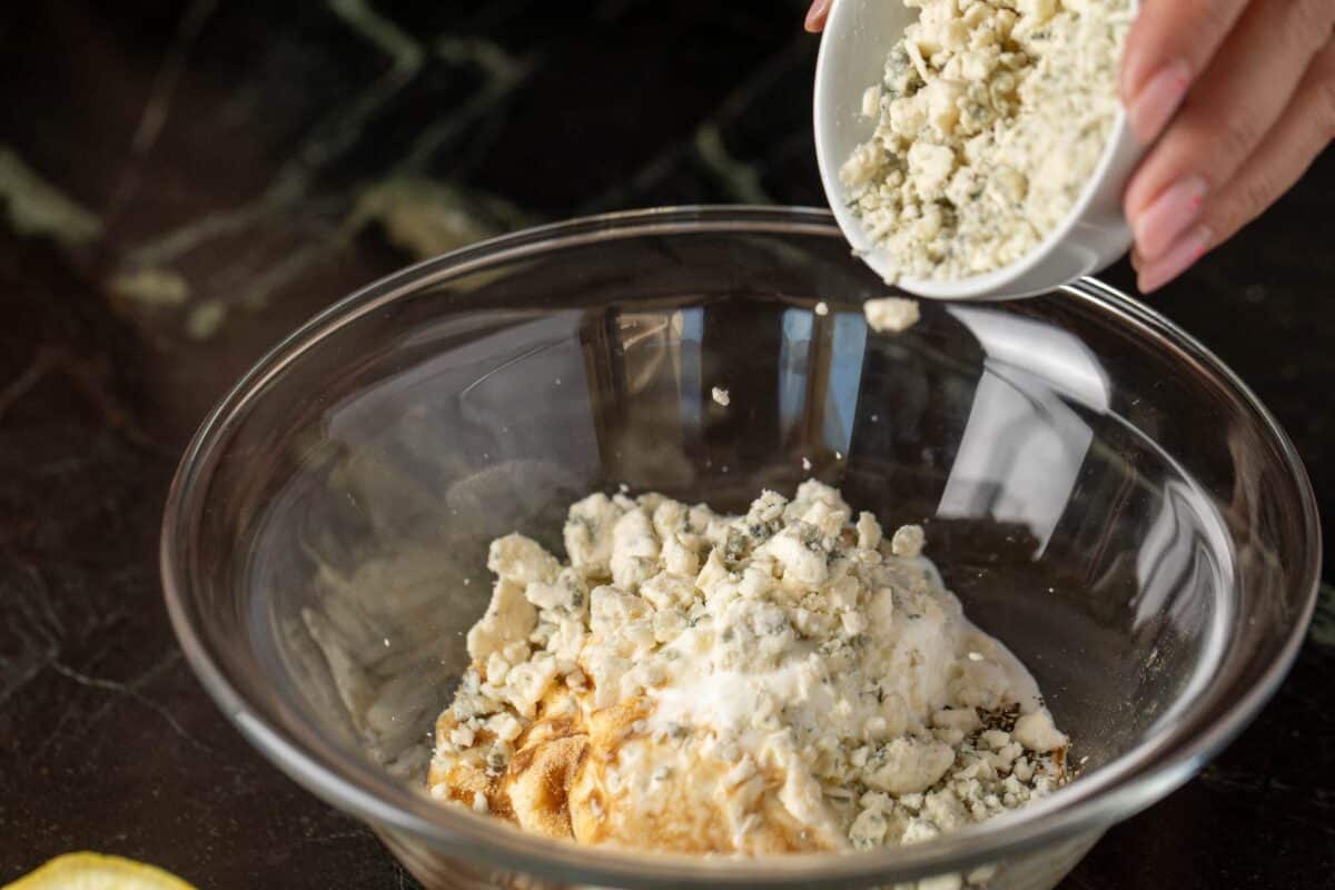 Blue cheese crumbles being added to ingredients in glass bowl.