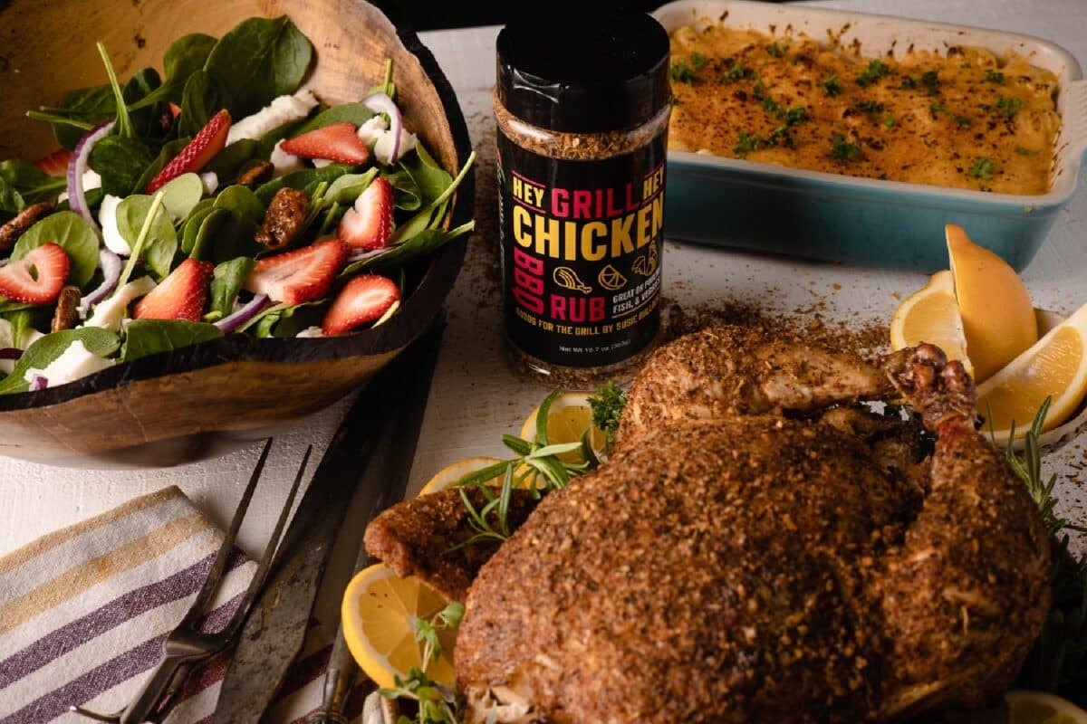 Bottle of Chicken Rub on table next to white dish with potatoes, platter of grilled chicken, and a bowl of salad.