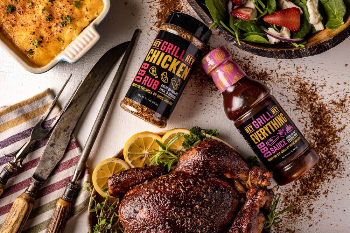 Bottles of Chicken Rub and Everything BBQ Sauce on tabletop next to tongs, and plates of grilled food.