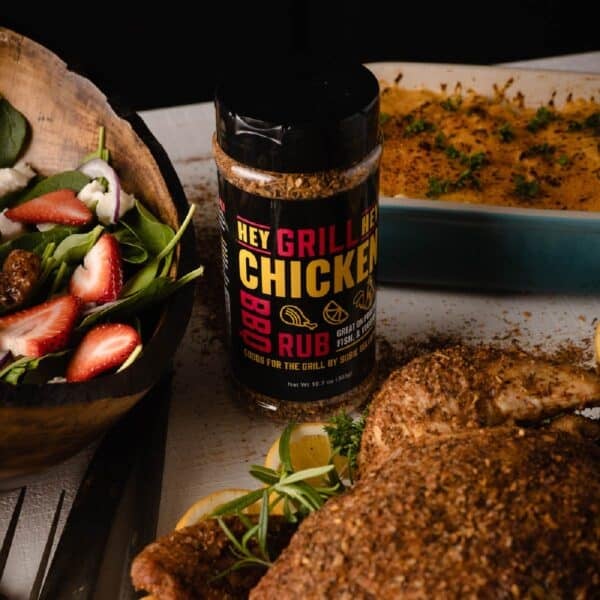 Bottle of Chicken Rub on table next to white dish with potatoes, platter of grilled chicken, and a bowl of salad.