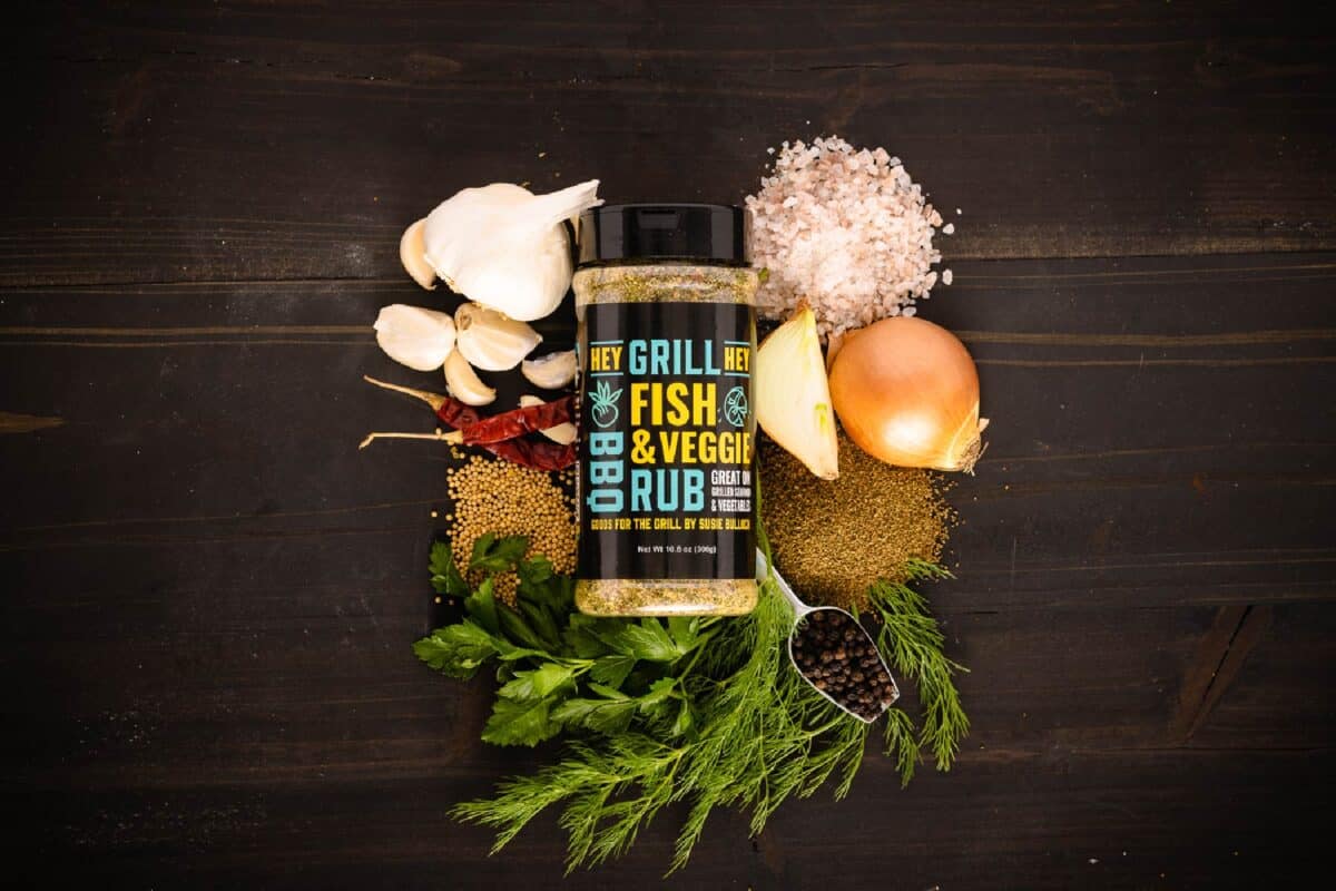 Jar of Fish & Veggie Rub surrounded by raw ingredients on black surface.