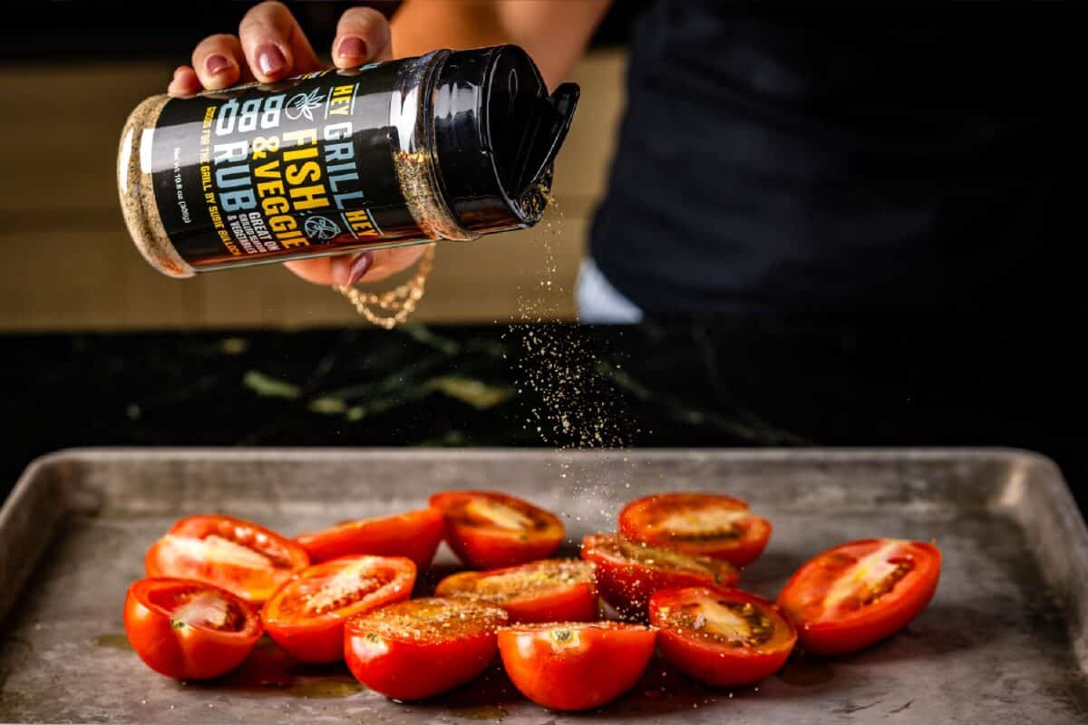 Sliced tomatoes on baking sheet being sprinkled with vegetable seasoning.