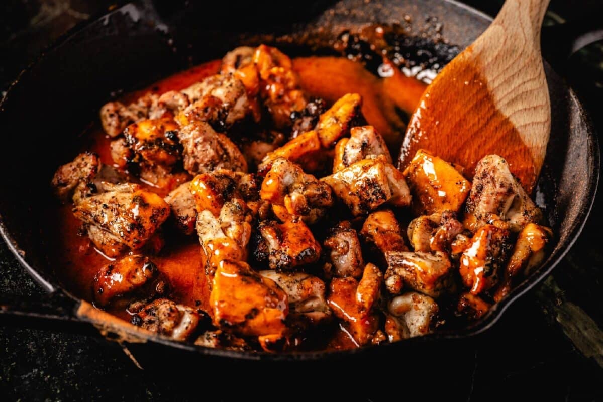 Chicken thighs being mixed into buffalo sauce in skillet with wooden spoon.