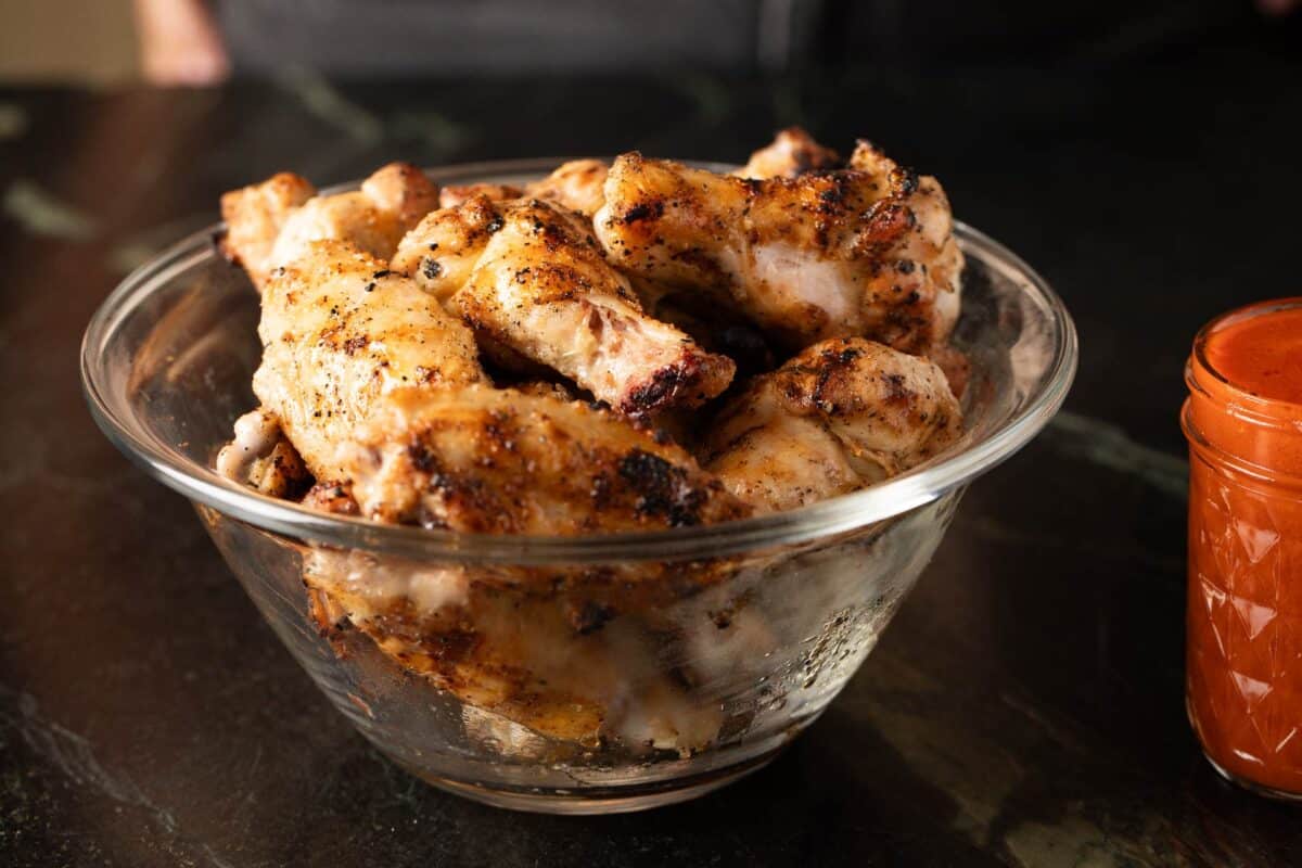 Chicken wings in glass bowl next to jar of buffalo sauce.