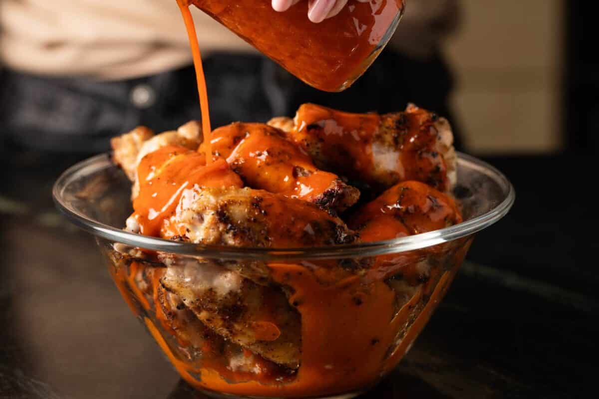 Buffalo sauce being poured onto wings in glass bowl.